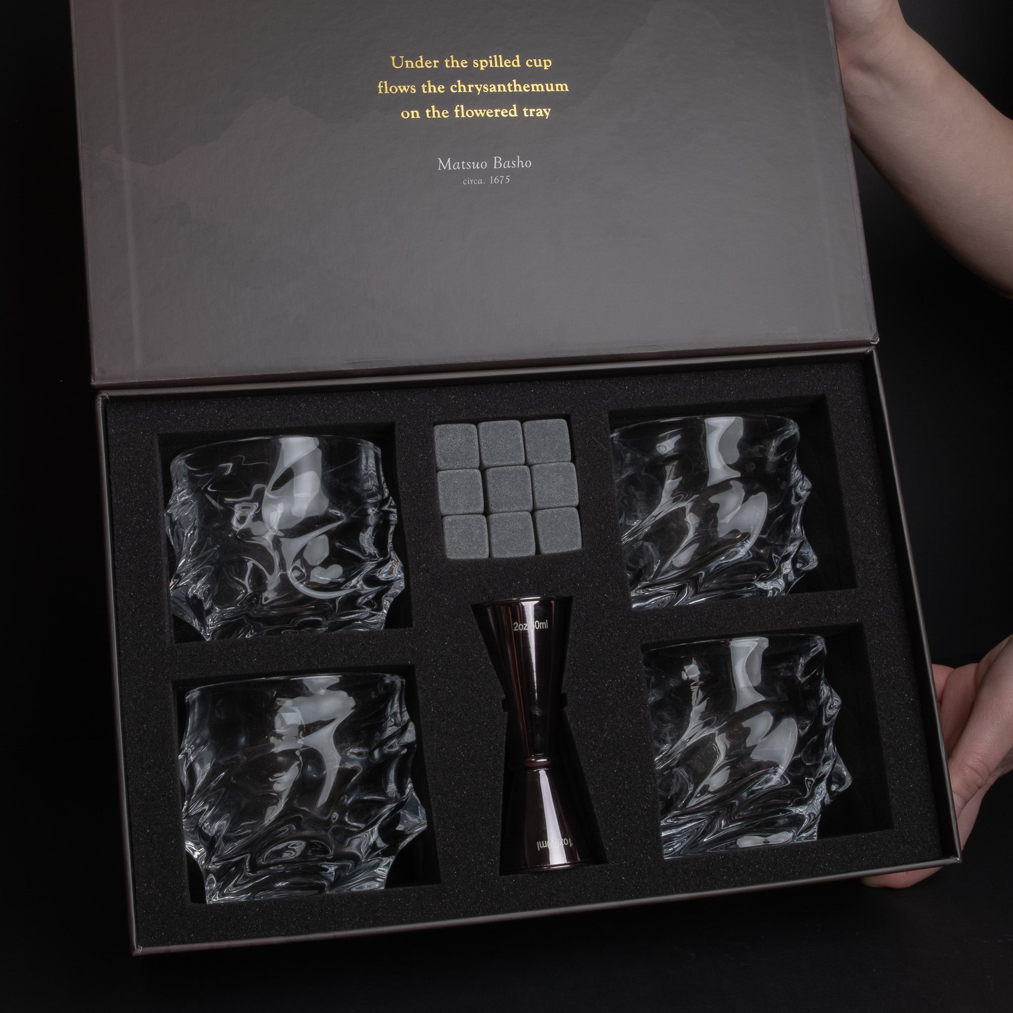 A premium crystal whiskey glass set in an open presentation box, featuring four textured clear crystal glasses, a central metallic jigger, and four gray ice cubes. The dark gray box lid has gold text and is held open by hands at the top right and bottom right.