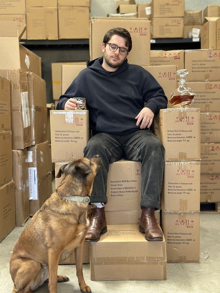 Thomas on a throne of stock boxes holding a decanter, Luna beside him, October 2025